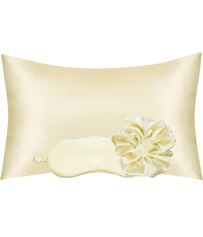 A beige satin pillowcase with a decorative flower detail at the bottom, includes a matching eye mask and scrunchie.